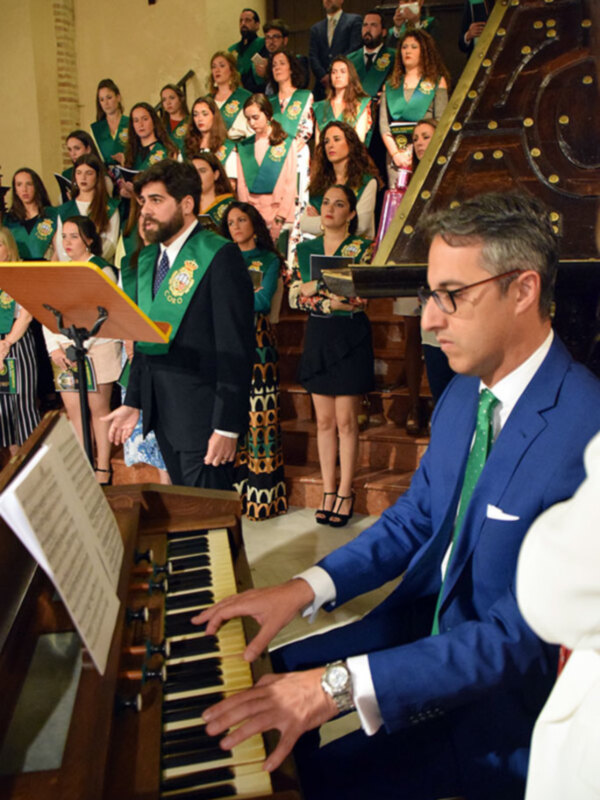 Un coro de jóvenes cantantes en trajes formales, con un hombre tocando el piano a su derecha. La escena se desarrolla en una sala de música con arcos y un órgano en el fondo.