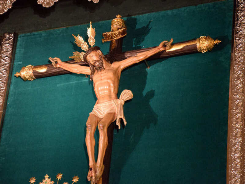 Una escultura de Jesucristo crucificado en un fondo verde. La figura está en posición vertical, con las manos extendidas y los pies conectados a la cruz. La escultura está dorada con detalles en oro, y se encuentra dentro de un marco de madera.