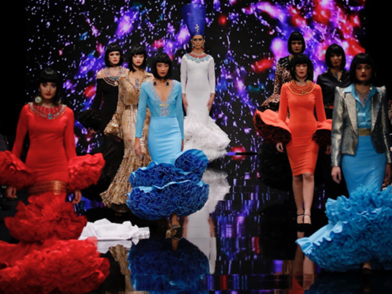 Modelos en un desfile de moda con vestidos de colores vibrantes y diseños llamativos. Fondo estrellado iluminado con luces de colores.