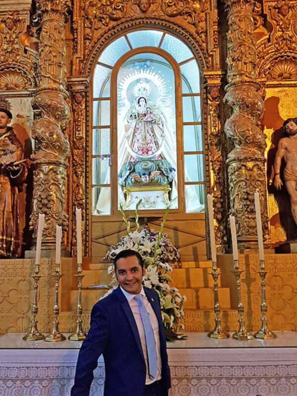 Se puede ver a una persona vestida con un traje formal, posando frente a un altar de una iglesia. El altar está decorado con estatuas y adornos dorados, y en el fondo se puede observar un arco de cristal que permite ver la luz del sol. La decoración y el ambiente sugieren una atmósfera religiosa y festiva.