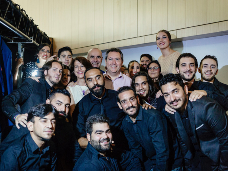 Una foto de grupo de personas sonriendo y posando en una sala con cortinas azules. La imagen muestra a varios hombres y mujeres vestidos de manera formal, posando juntos frente a una pantalla. La escena parece ser en un evento o reunión formal.