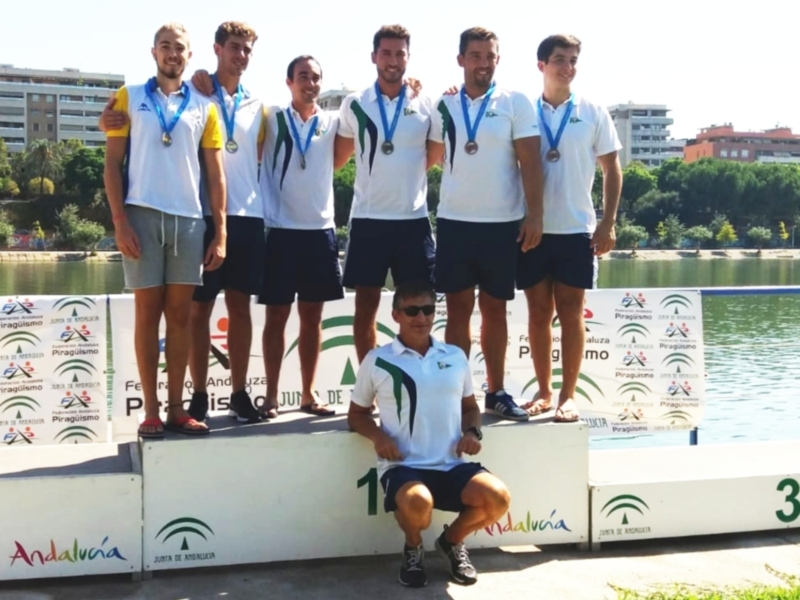 Un grupo de cinco hombres posa en un podio con medallas, celebrando una victoria deportiva. El hombre en el centro está sentado y sonríe, mientras los demás están de pie detrás de él. El fondo muestra un paisaje urbano con edificios y árboles, junto a una zona de agua. La imagen está firmada con "Andalucía".