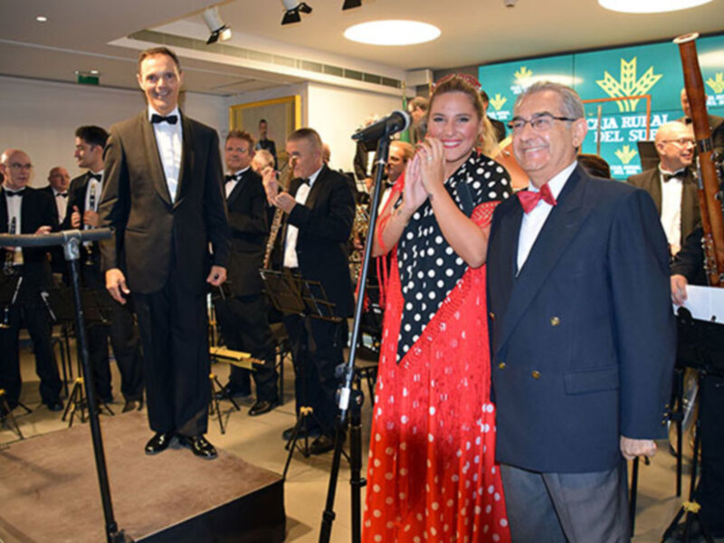 Una imagen de una gala o evento formal donde varios hombres y mujeres están vestidos con trajes elegantes. La escena se centra en una pareja principal, un hombre y una mujer, que están sonriendo y saludando. El hombre lleva un traje negro con corbata, mientras que la mujer está en un vestido rojo y blanco con puntos blancos. En el fondo, hay otros asistentes que también están vestidos formalmente. La iluminación es brillante, y la decoración sugiere un ambiente festivo o ceremonial.