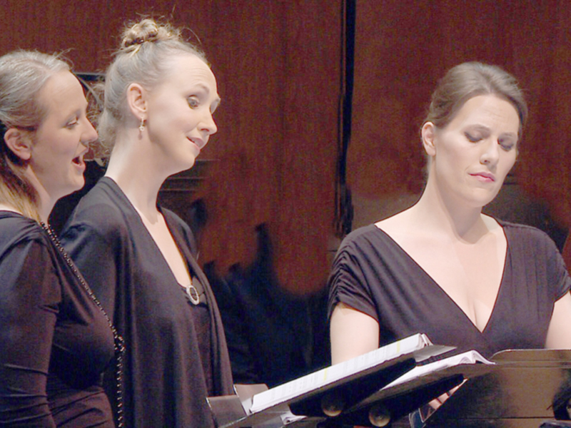 Tres mujeres en un escenario, posiblemente cantantes o actrices, sonríen y miran hacia la derecha. La imagen sugiere una escena de la vida en el teatro o concierto, con un fondo rojo que podría ser una cortina.