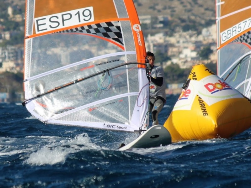 Vela de windsurf con bandera española y número de competición ESP19, navegando en el mar con una ciudad en el fondo.