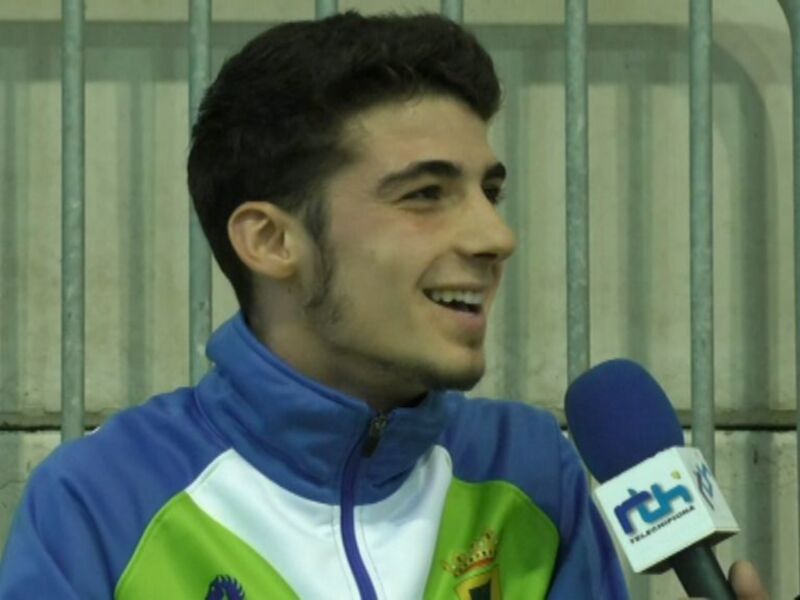 Un hombre con una chaqueta deportiva azul y verde, sonriendo mientras habla en un micrófono con la etiqueta "RTH". Fondo de una estructura metálica.