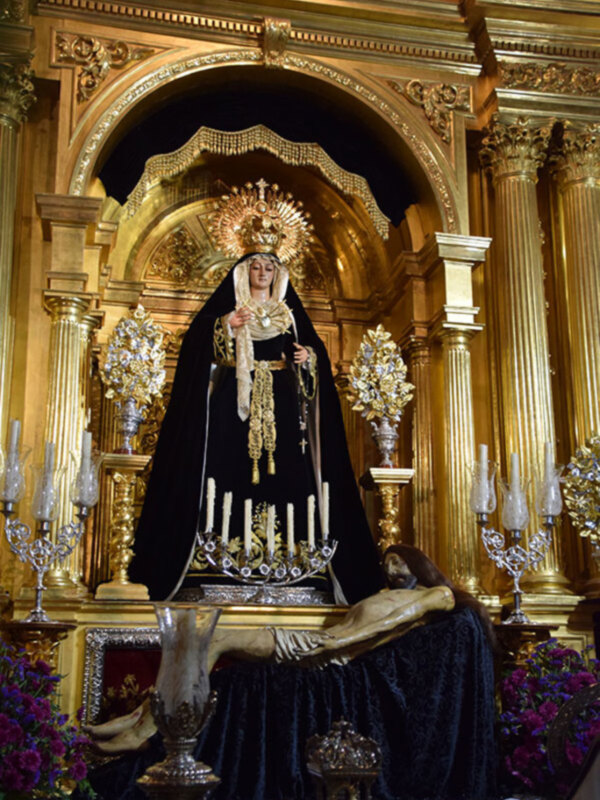 Imagen de una Virgen María vestida con un manto negro y coronada, sentada en un altar dorado. A su lado, se encuentra una estatua de Cristo crucificado. La escena está rodeada por flores y adornos dorados, en un ambiente de lujo y solemnidad.