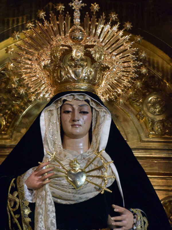 Imagen de la Virgen María con un halo radiante y una corona dorada, vestida con un manto negro y una saya dorada. La imagen se encuentra en un entorno arquitectónico con detalles dorados y una estructura de madera.