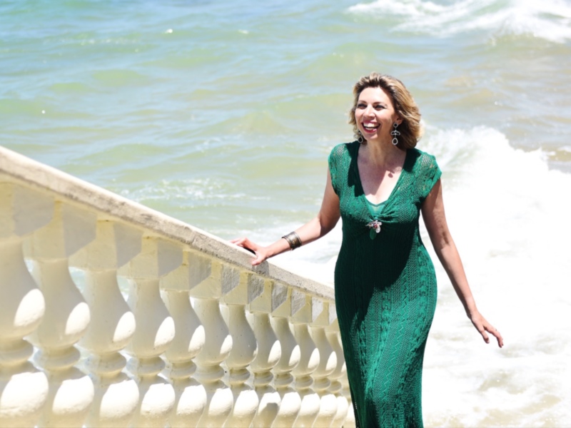 Una mujer en un vestido verde brillante posa frente al mar, con olas y una balaustrada de piedra detrás.