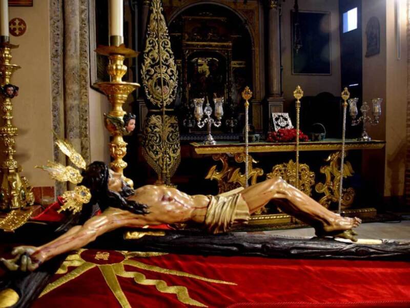 Representación del Cristo de la Buena Muerte en una procesión religiosa, con trono dorado y candelabros, en un entorno eclesiástico.