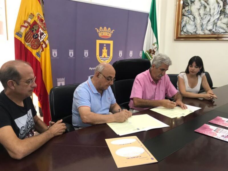Tres personas firmando documentos en una sala con banderas y un escudo de armas en el fondo.