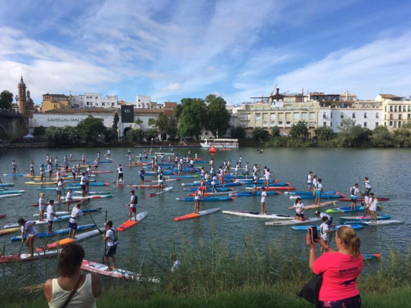 Una multitud de personas disfruta del deporte de surfboard en un lago, con edificios y árboles al fondo. Un hombre toma una foto de la escena, mientras que otra persona disfruta del paisaje.
