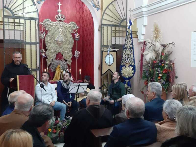 Una celebración religiosa en una iglesia con una imagen de la Virgen en el altar, mientras un hombre habla al micrófono y otros participan en la música.