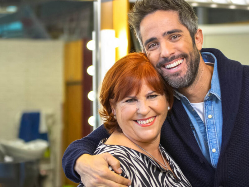 Dos personas sonrientes, uno con cabello rojo y la otra con cabello corto. El ambiente parece ser un estudio de televisión o una producción de entretenimiento, con luces y decoración en el fondo.