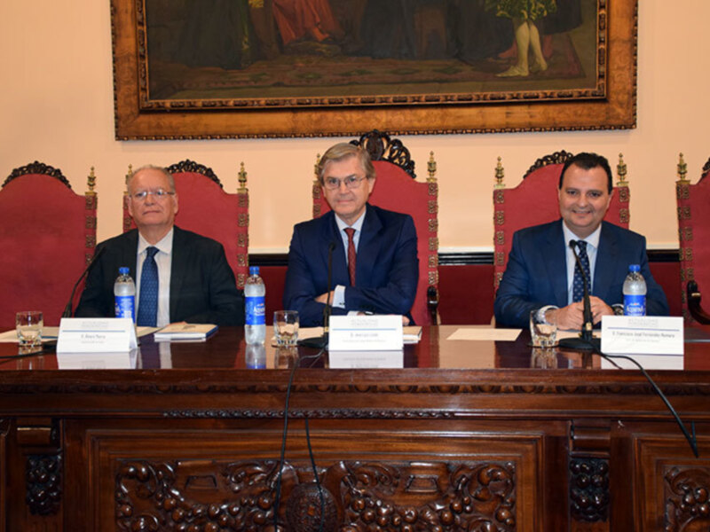 Tres hombres sentados en una sala formal, con micros en el escritorio y una imagen histórica en el fondo. La escena sugiere una reunión oficial o una conferencia de prensa.