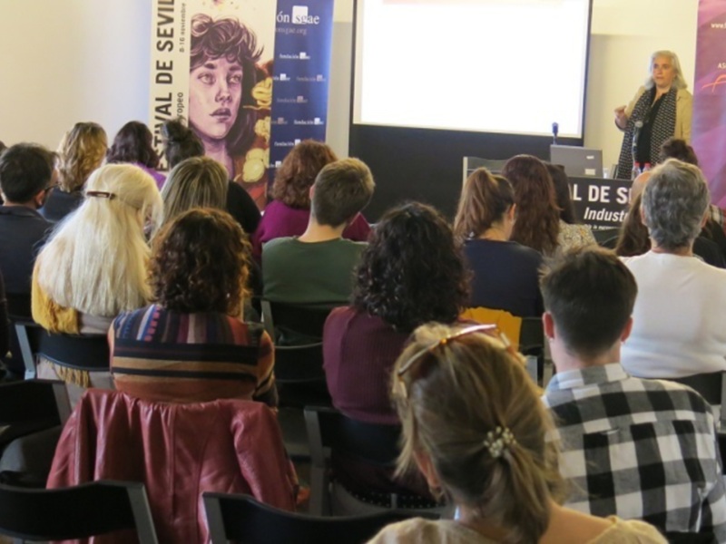 Una audiencia en una sala de conferencias, con carteles promocionales y una ponente en el escenario.