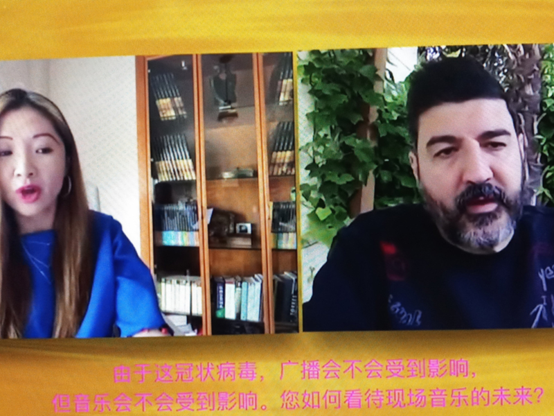 Dos personas en una videoconferencia, uno con cabello largo y una barba, frente a un fondo de estanterías llenas de libros. Texto en chino abajo.