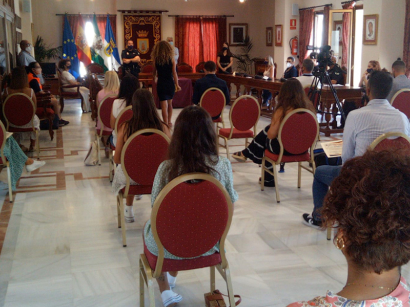 Una sala de conferencias con personas sentadas en sillas rojas, una bandera europea y un escudo de armas en el fondo.