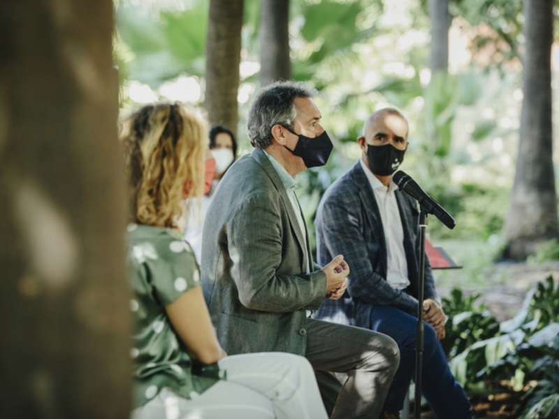 Una imagen de tres personas en una reunión al aire libre, con árboles y vegetación al fondo. La persona central está hablando en un micrófono, mientras que las otras dos escuchan.