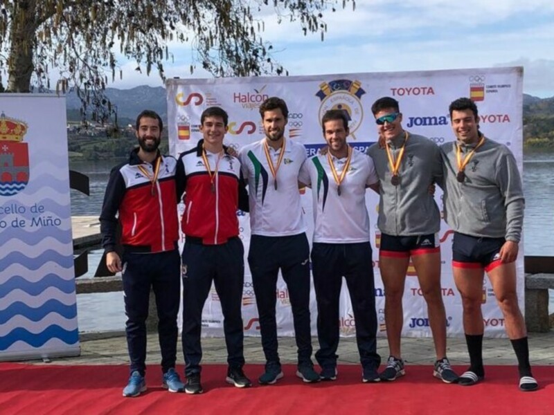 Ganadores de una competición de remo posan en el podio, con banderas de España y patrocinadores en el fondo.