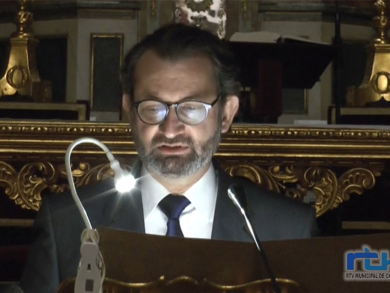 El hombre está hablando en un escenario formal, con una iluminación que enfatiza su rostro y la estructura histórica al fondo.