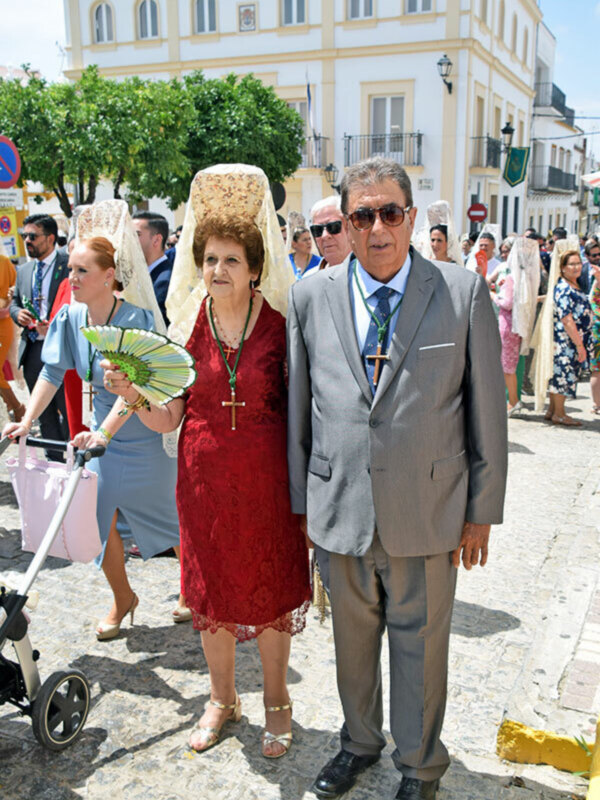 Un hombre y una mujer en traje caminan por la calle, con el hombre usando gafas de sol y la mujer llevando un vestido rojo y una saya. La escena parece ser en un entorno urbano con edificios y gente a su alrededor.