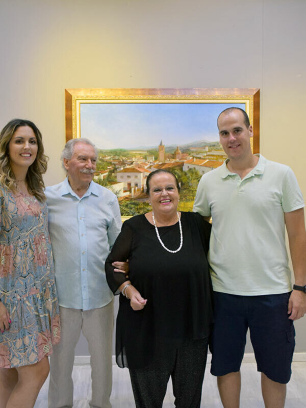 Una familia de cuatro personas posa frente a una pintura que representa un paisaje con edificios y montañas. La imagen muestra a una mujer en el centro, dos hombres de ambos lados y una joven a la izquierda. Todos parecen estar en una sala con un suelo de mármol y paredes blancas.