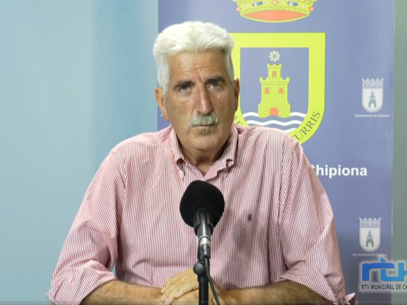 El hombre está en una conferencia de prensa, frente a un fondo azul con el escudo municipal y la bandera de Chipiona. Está hablando en un micrófono, con una camisa rosa a cuadros y cabello blanco.