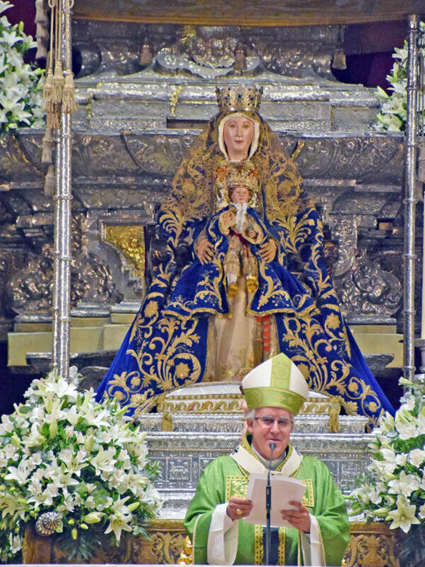 Un sacerdote vestido con un hábito verde y una saya blanca, con un cíngulo dorado, está leyendo un libro en el altar. En la parte superior del altar, hay una imagen de una Virgen María vestida con un manto azul y dorado, sosteniendo a un niño. El altar está decorado con flores blancas y doradas, y hay una estructura metálica en el fondo.