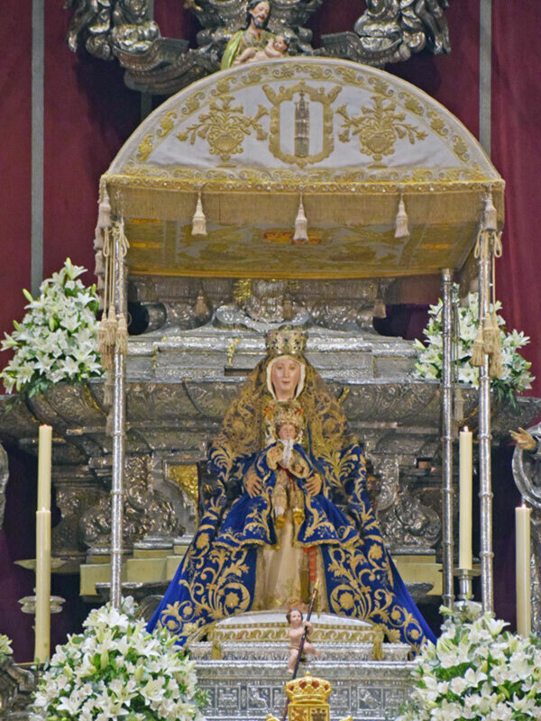Imagen de una Virgen María vestida en trajes dorados y azules, sentada sobre un altar decorado con flores blancas. El fondo es de color rojo y hay detalles dorados en la parte superior del altar.