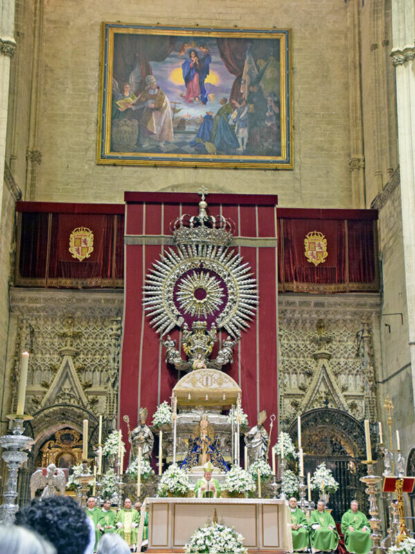 Interior de una iglesia con un altar grande adornado con flores y velas, y una imagen de la Virgen en el centro. En el fondo, hay un lienzo con una escena religiosa.