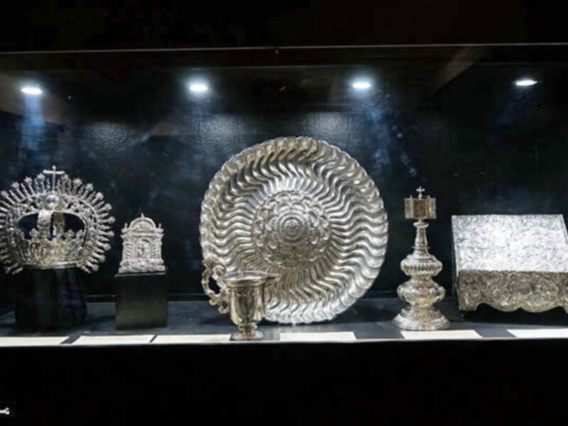 Un conjunto de objetos de plata, incluyendo una corona, un plato con forma de concha, una lámpara y un ataúd, es exhibido en un museo. La iluminación destaca los detalles y la belleza de estos objetos históricos.