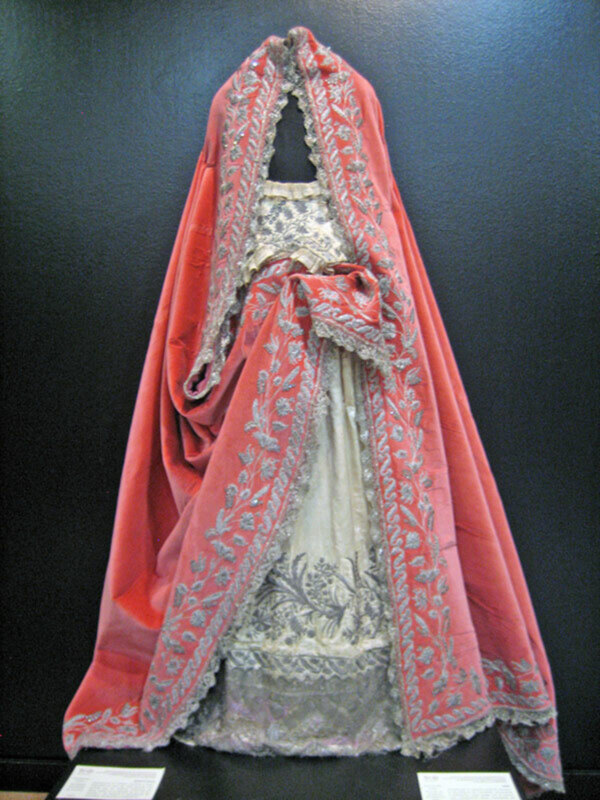 Una imagen de un vestido tradicional con un capote rojo y bordado en beige, colgando frente a una pared azul.