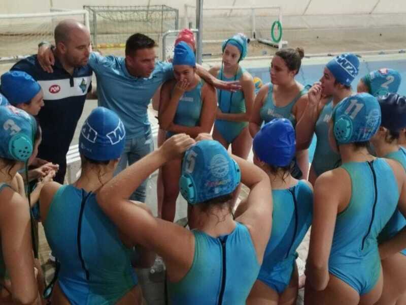 Equipo de natación femenina en un entrenamiento, con nadadoras vestidas con trajes azules y gorros de natación. Un entrenador observa a los nadadores en la piscina.