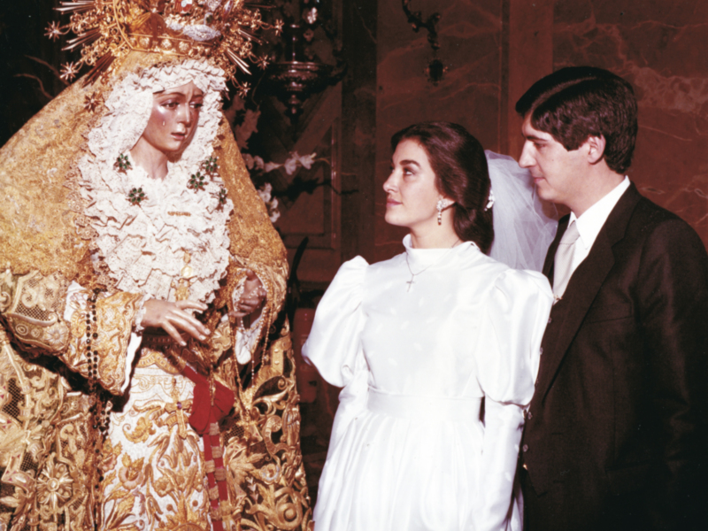 Imagen de una mujer vestida con un traje tradicional y una corona, posiblemente en una celebración religiosa o cultural. A su lado, dos personas más están mirando hacia la izquierda. La escena parece ser en un entorno interior, posiblemente una iglesia o sala de eventos.