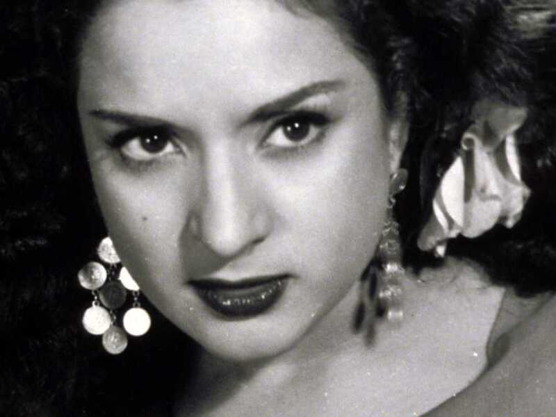 A una mujer con cabello largo y ondulado, posiblemente en un estilo vintage. Se pueden ver detalles como su expresión facial serena y sus ojos grandes, así como sus joyas, incluyendo pendientes y un collar. La imagen es en blanco y negro, lo que añade un tono nostálgico o retro al retrato.
