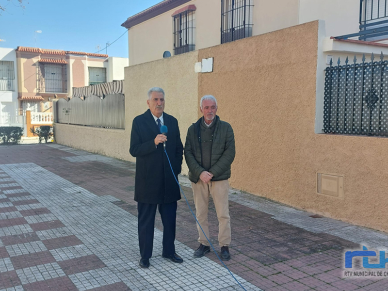 Dos hombres se encuentran en una calle con adoquines cuadriculados. El hombre de la izquierda, vestido con traje y corbata, sostiene un micrófono. El hombre de la derecha, con chaqueta verde y pantalones beige, sostiene una vara de lectura. Fondo: casas con tejas rojas y azules, muro beige con ventanales.