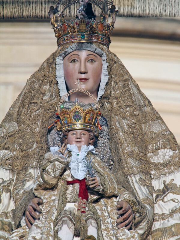 Estatua de la Virgen María con una corona y vestimenta elaborada, sosteniendo un niño. Contexto religioso o devocional.