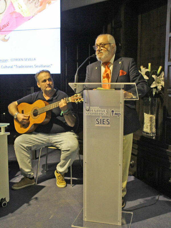 Un hombre toca la guitarra mientras otro habla en un podium con el logo de SIES. La imagen muestra una presentación o conferencia sobre tradiciones suizas, con un fondo de pantalla que incluye información sobre Citroen Sevilla.