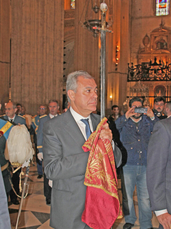 Imagen de un hombre en traje sosteniendo una bandera roja con detalles dorados, posiblemente en un evento formal o ceremonial. El fondo muestra una iglesia con vitrales y estructuras arquitectónicas.