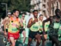 Corredores participando en una carrera de maratón urbana bajo un cielo azul claro, con edificios y árboles en el fondo.