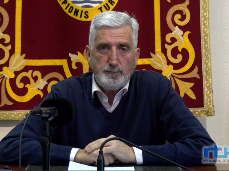 Un hombre con barba gris y cabello blanco, sentado en una mesa con un fondo de bandera roja y dorada.