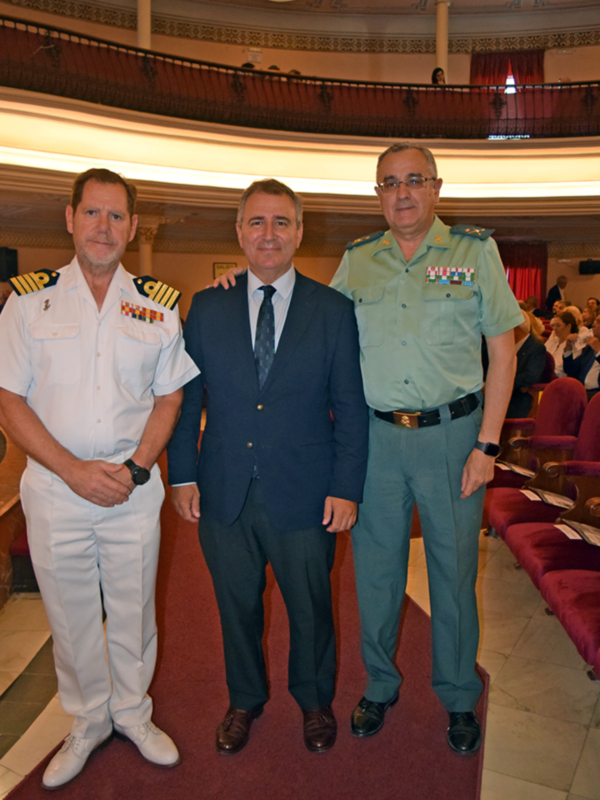 Tres hombres en uniforme de oficiales militares posan juntos en un salón con arcos y luces. El hombre izquierdo lleva uniforme blanco, el centro un traje azul y el derecho una camisa verde con insignias militares. La imagen se centra en su gesto de camaradería y uniforme distintivo.