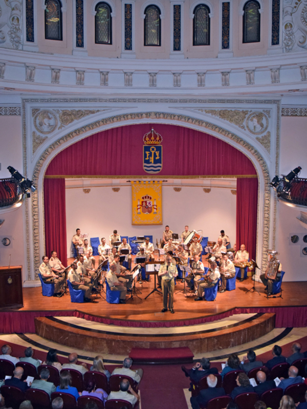 Un concierto en un teatro con una orquesta en el escenario y un público sentado. La imagen muestra a músicos tocando instrumentos de viento, cuerda y percusión. La orquesta está en el centro del escenario, con un director frente a ellos. En la parte superior de la imagen se puede ver una bandera con un escudo y una corona. La iluminación es adecuada para el concierto, con focos en las personas y la orquesta.