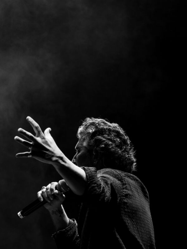 Cantante en escenario con mano levantada, iluminación dramática y humo en el fondo.