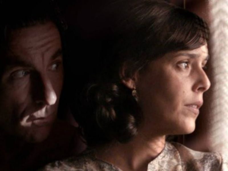 Dos personas mirando hacia la izquierda, uno de ellos con una expresión seria y el otro con un semblante pensativo. La escena parece ser interior, posiblemente en una habitación con cortinas de fondo.