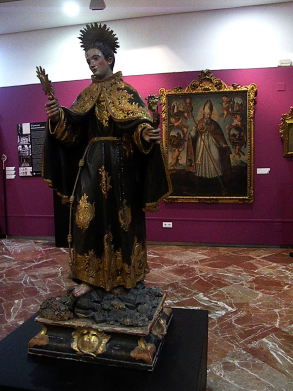 Elegante estatua religiosa con vestimenta dorada y negra, posiblemente de un santo, en un museo con fondo de pintura y piso de mármol.