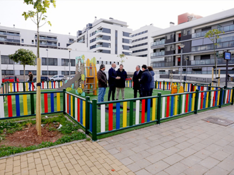 Un grupo de personas observa una escuela infantil con un jardín verde y un árbol recién plantado. El área está rodeada por una cerca de madera pintada con colores brillantes. En el fondo, se pueden ver edificios blancos y modernos de varios pisos.