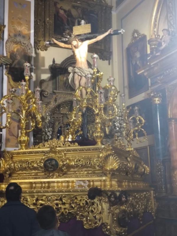 Se puede ver una estatua dorada de un personaje crucificado, probablemente Cristo, en el centro de una estructura elaborada y ornamentada. A su alrededor hay varios cuadros y estatuas religiosas, creando un ambiente de iglesia. La iluminación es intensa, enfocándose en la estatua central.