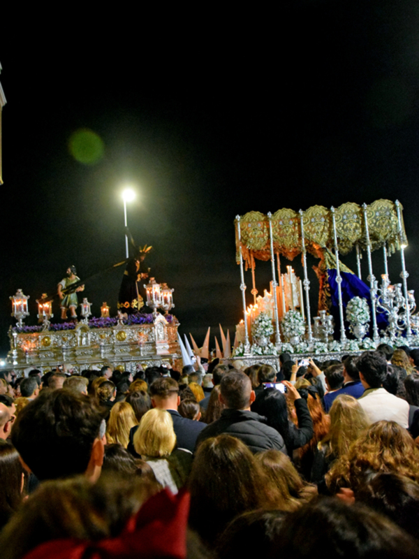 Una procesión nocturna con una imagen de Cristo en el centro, rodeada por faroles y banderas, mientras la multitud observa desde abajo.